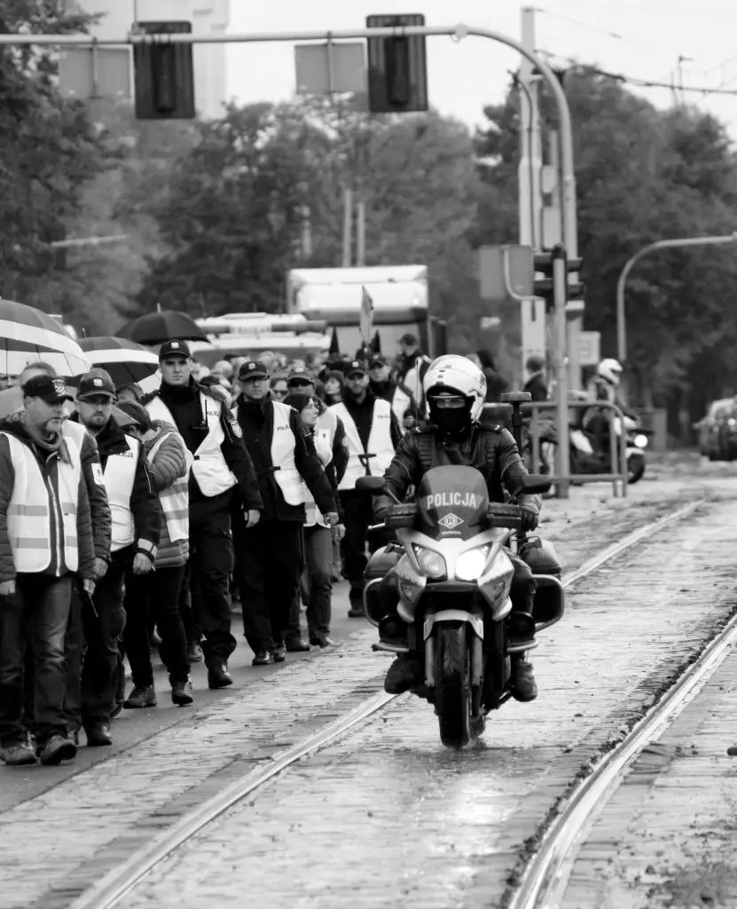 Black and white city parade with police motorbike leading march through wet street.