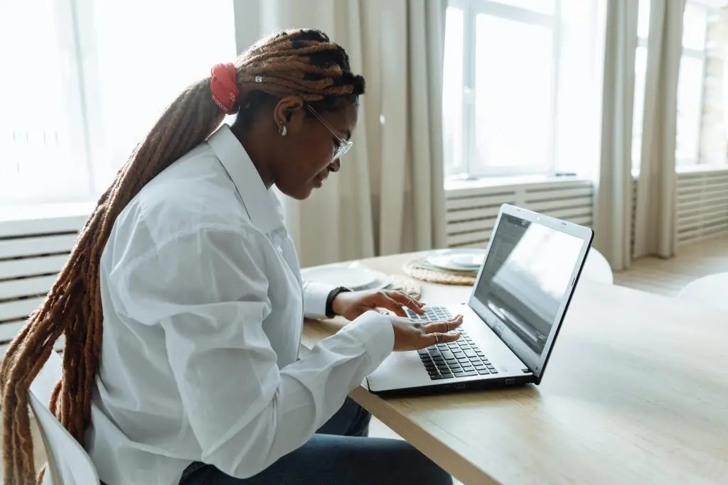 A woman with dreadlocks focuses intently on her laptop in a bright, cozy home setting.