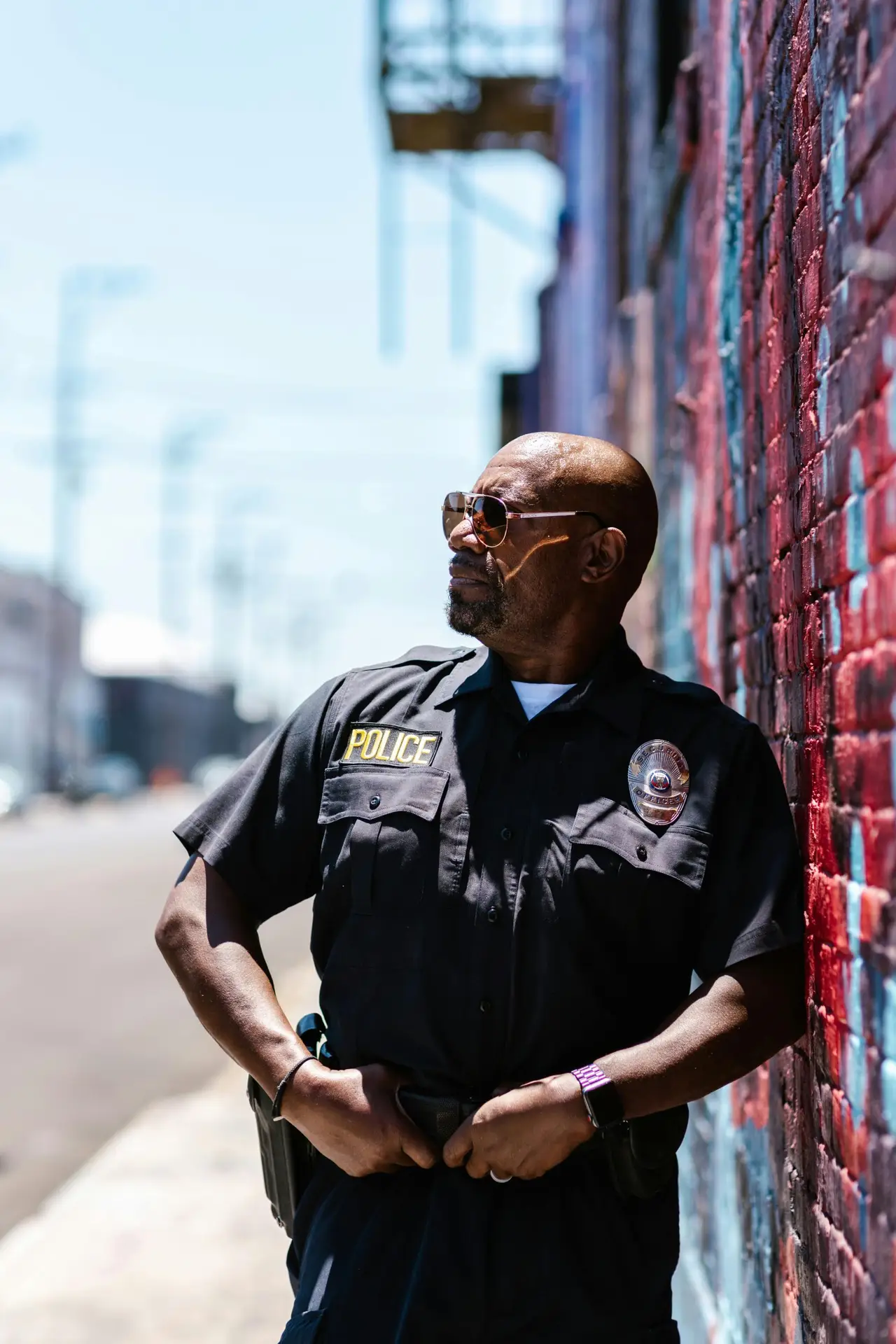 Bald African American police officer in uniform standing against a painted wall outdoors.